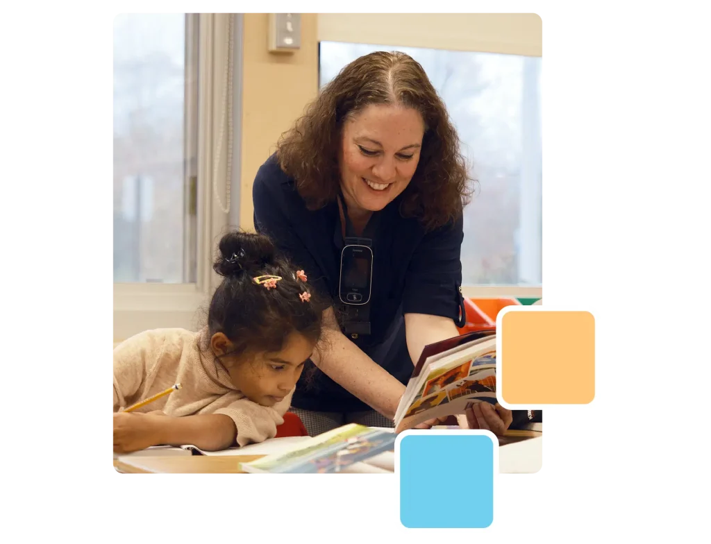 An adult helps a child read a book at a table. The child is writing, and the adult is smiling. The setting resembles a classroom, with windows in the background.